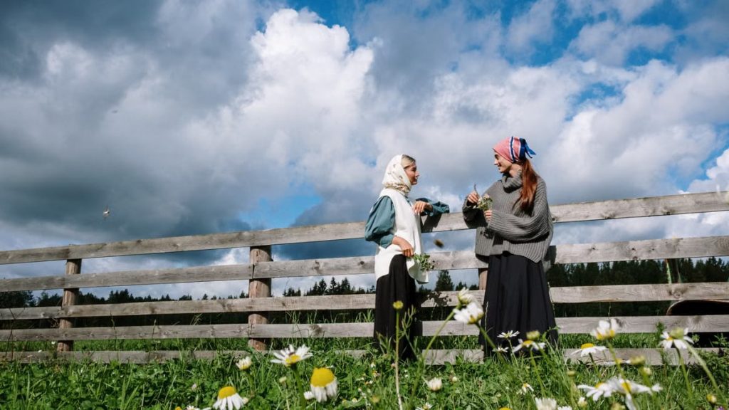 Two women in headscarves enjoying a conversation by a rustic fence in a flower-filled meadow.