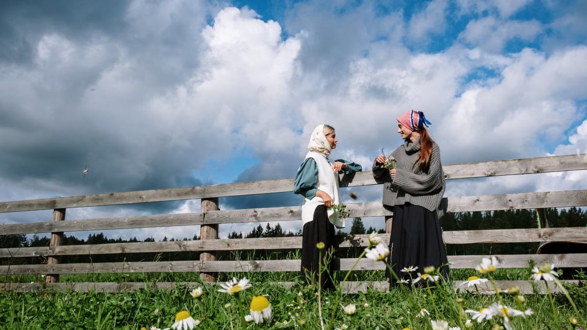 Two women in headscarves enjoying a conversation by a rustic fence in a flower-filled meadow.